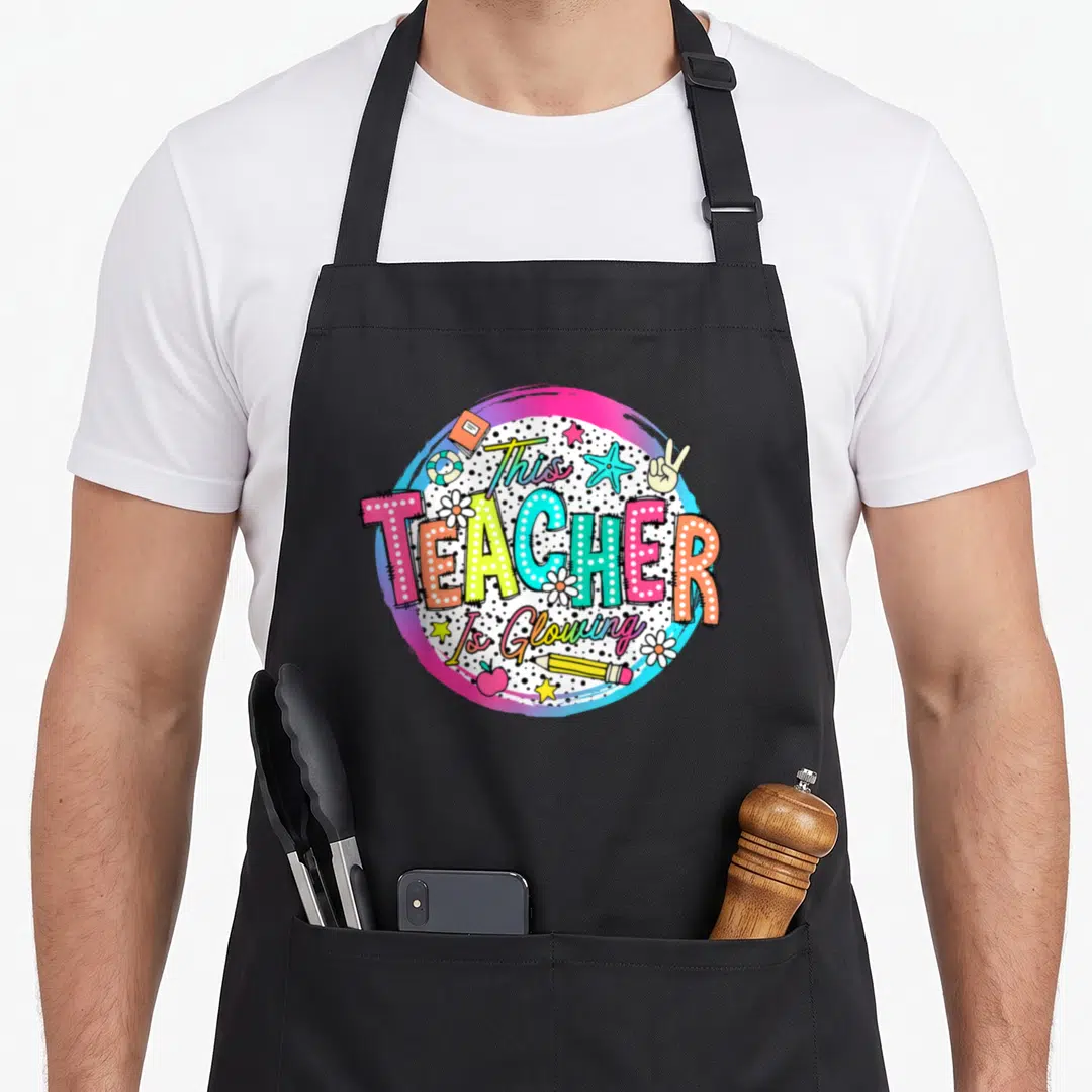 Glowing teacher apron - This TEACHER Is Glowing text with summer icons inside polka dot circle — shown on a black cotton canvas apron against a white background