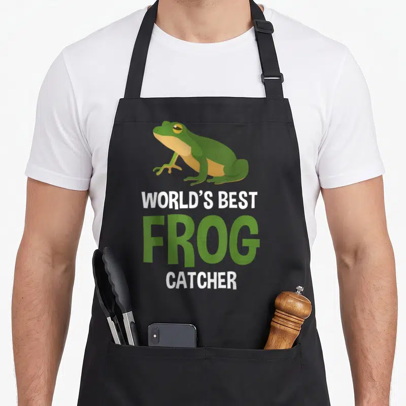Catcher frog aprons - Green cartoon frog above white and green text reading WORLD'S BEST FROG CATCHER — shown on a black cotton canvas apron against a white background