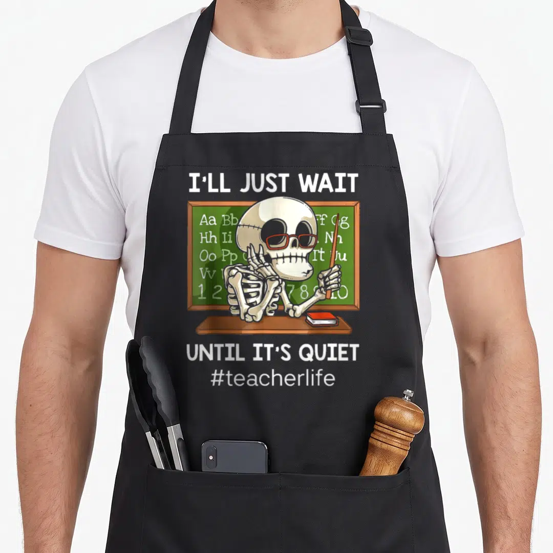 Wait teacher apron - Skeleton with red glasses leaning on desk in front of green chalkboard with alphabet and numbers — shown on a black cotton canvas apron against a white background