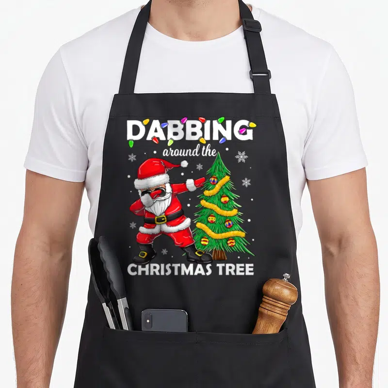 Around christmas tree apron - Cartoon Santa dabbing pose next to green Christmas tree with colorful ornaments — shown on a black cotton canvas apron against a white background