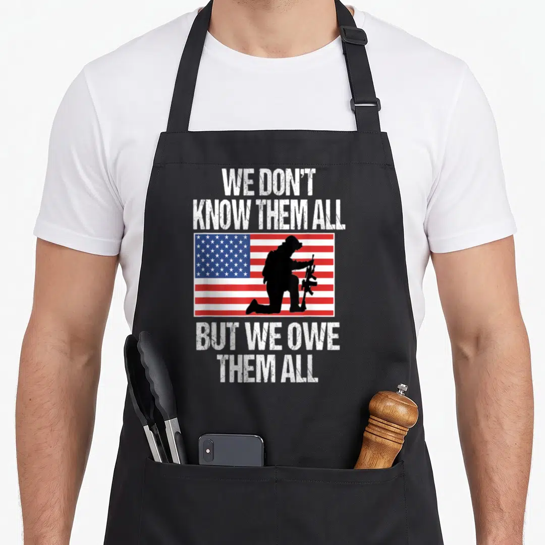 Them memorial apron - Kneeling soldier silhouette on USA flag with WE DON'T KNOW THEM ALL BUT WE OWE THEM ALL text — shown on a black cotton canvas apron against a white background