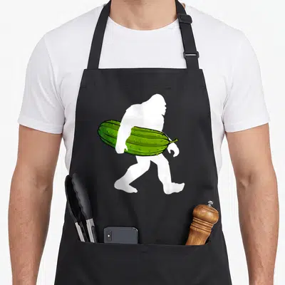 Bigfoot pickle apron - White bigfoot silhouette carrying a bumpy green cucumber under one arm — shown on a black cotton canvas apron against a white background