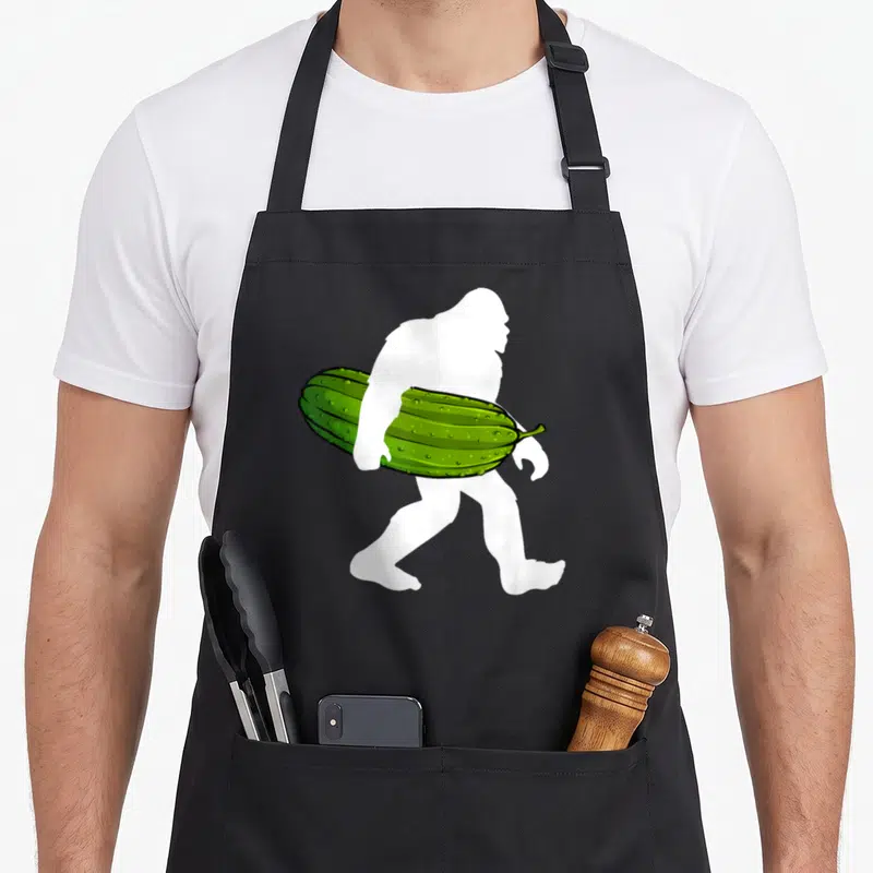 Bigfoot pickle apron - White bigfoot silhouette carrying a bumpy green cucumber under one arm — shown on a black cotton canvas apron against a white background