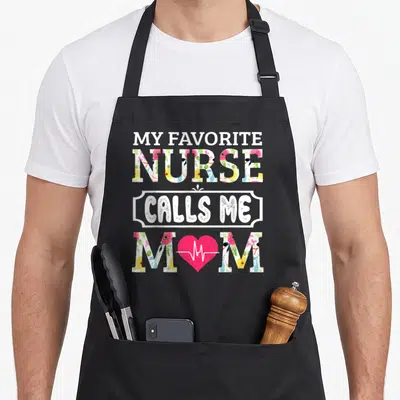 Vintage call white nurse apron - Floral typography reading My Favorite Nurse Calls Me Mom with pink heartbeat heart — shown on a black cotton canvas apron against a white background