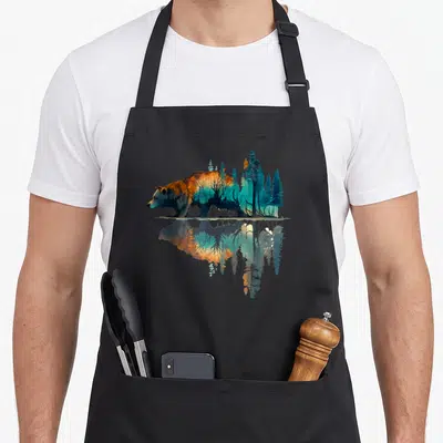 Reflection bear apron - Bear silhouette filled with blue and orange forest trees mirrored in water reflection — shown on a black cotton canvas apron against a white background