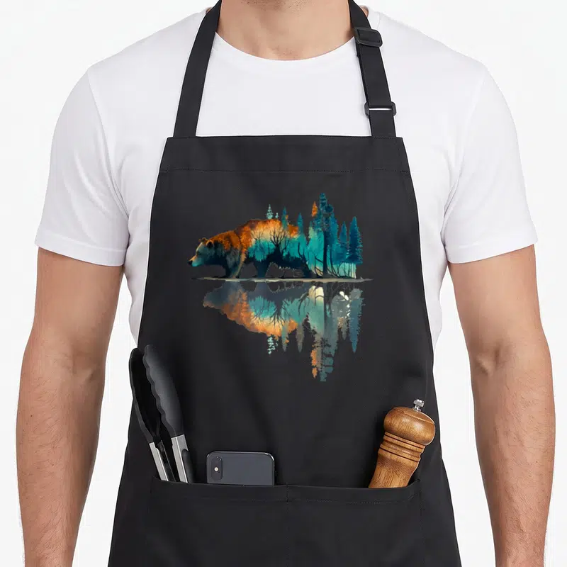 Reflection bear apron - Bear silhouette filled with blue and orange forest trees mirrored in water reflection — shown on a black cotton canvas apron against a white background