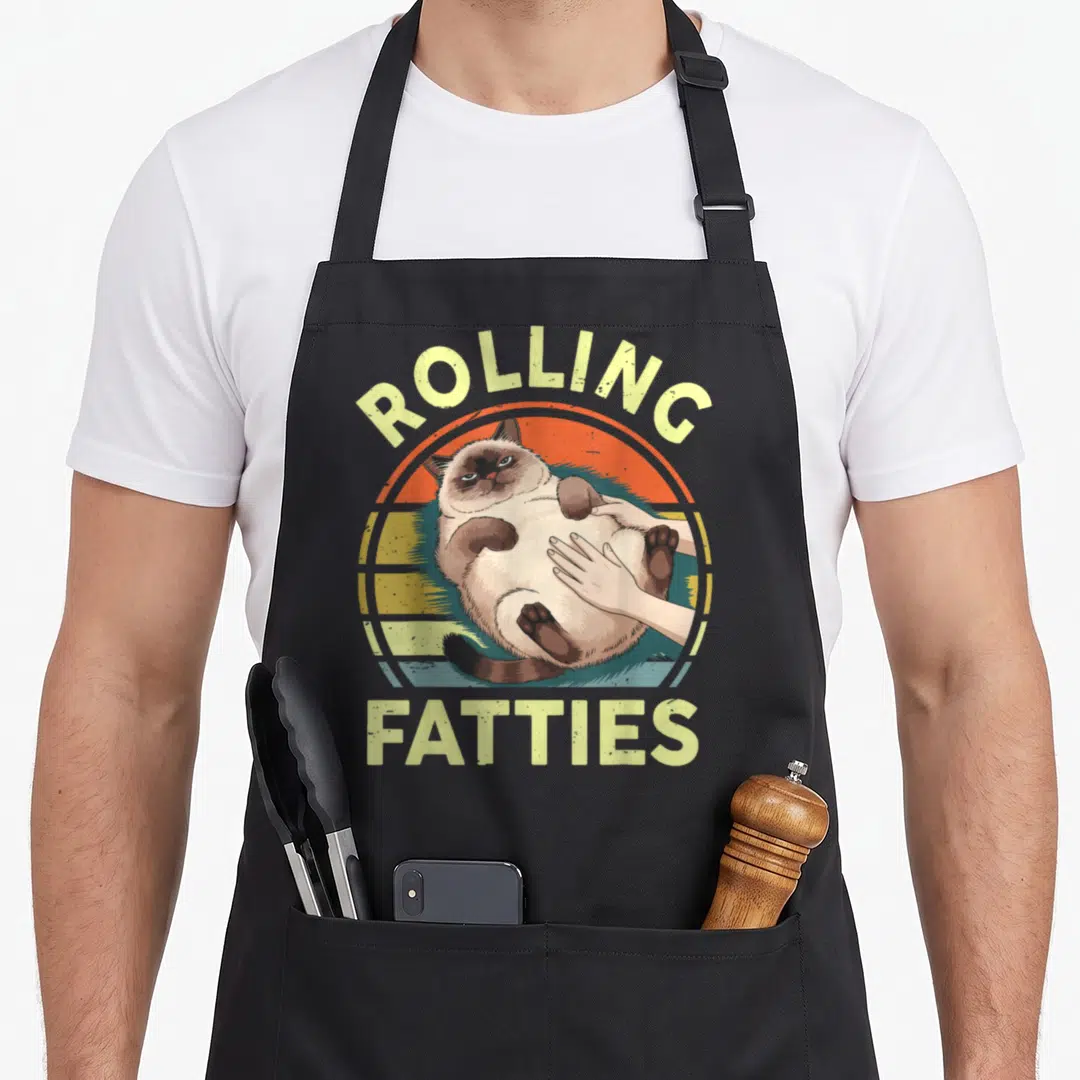 Chonky cute cat aprons - Fat cat lying on back with hand rubbing belly over striped sunset, text rolling fatties — shown on a black cotton canvas apron against a white background