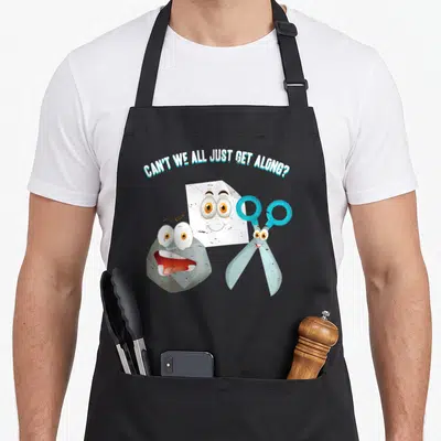 Along paper apron - Cartoon rock paper scissors characters with arched text Can't We All Just Get Along — shown on a black cotton canvas apron against a white background