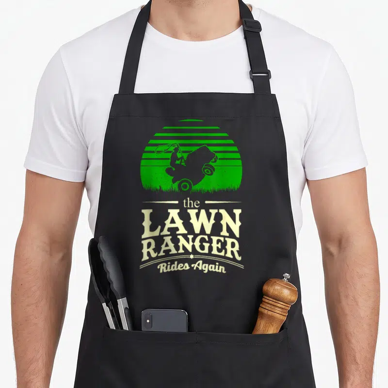 Grass rangers apron - Green silhouette of person on mower against striped sun with text The Lawn Ranger Rides Again — shown on a black cotton canvas apron against a white background