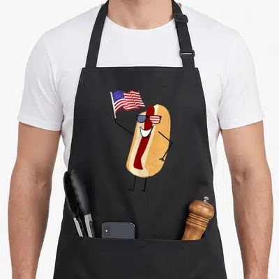 Picnic hot dog apron - Cartoon hot dog wearing flag sunglasses and holding a waving US flag — shown on a black cotton canvas apron against a white background
