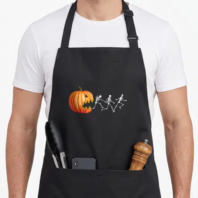 Pumpkin skeleton halloween apron - Orange pumpkin with jagged mouth chasing three white skeletons running rightward — shown on a black cotton canvas apron against a white background