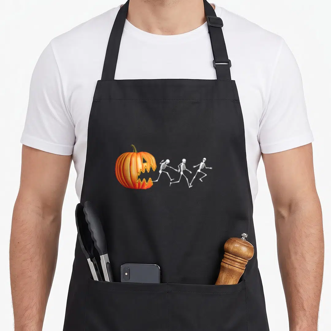Pumpkin skeleton halloween apron - Orange pumpkin with jagged mouth chasing three white skeletons running rightward — shown on a black cotton canvas apron against a white background