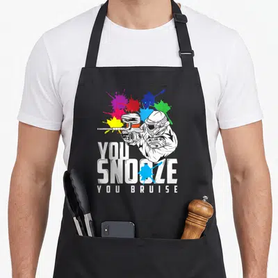 Bruise paintball apron - You Snoze You Bruise text with paintball player and blue red yellow splats — shown on a black cotton canvas apron against a white background