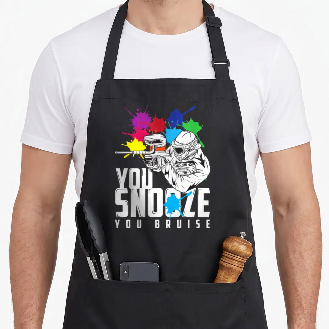 Bruise paintball apron - You Snoze You Bruise text with paintball player and blue red yellow splats — shown on a black cotton canvas apron against a white background
