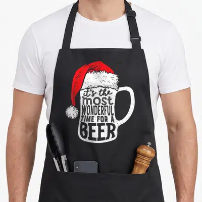 Wonderful santa aprons - White beer mug wearing red Santa hat with distressed text reading it's the most wonderful time — shown on a black cotton canvas apron against a white background