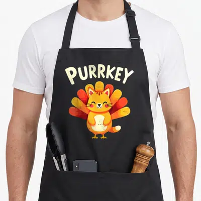 Purrkey funny turkey apron - Cartoon orange cat with red and yellow tail feathers under arched PURRKEY text — shown on a black cotton canvas apron against a white background