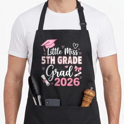 Graduation miss grade apron - Pink graduation cap, floral 5TH GRADE text, bow, and diplomas stacked centrally — shown on a black cotton canvas apron against a white background