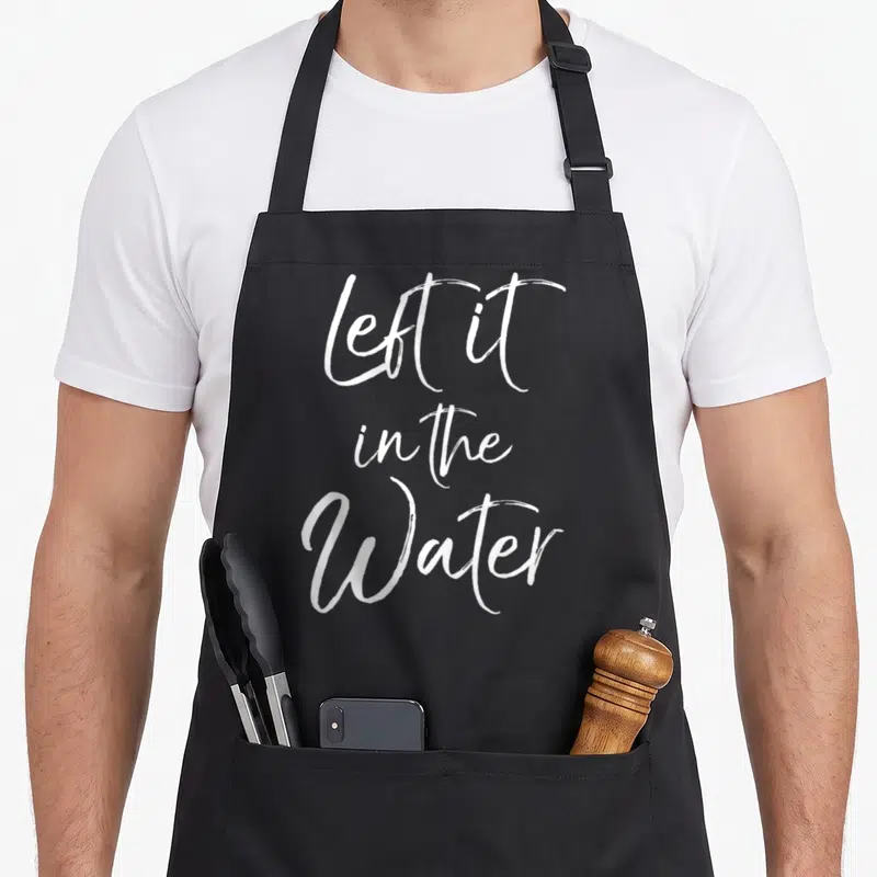 Left water christian apron - White cursive text left it in the Water stacked vertically on grey background — shown on a black cotton canvas apron against a white background