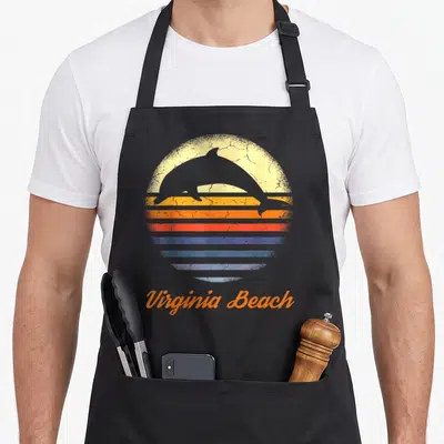 Virginia beach lover apron - Retro sunset with leaping dolphin silhouette and curved Virginia Beach text in orange — shown on a black cotton canvas apron against a white background