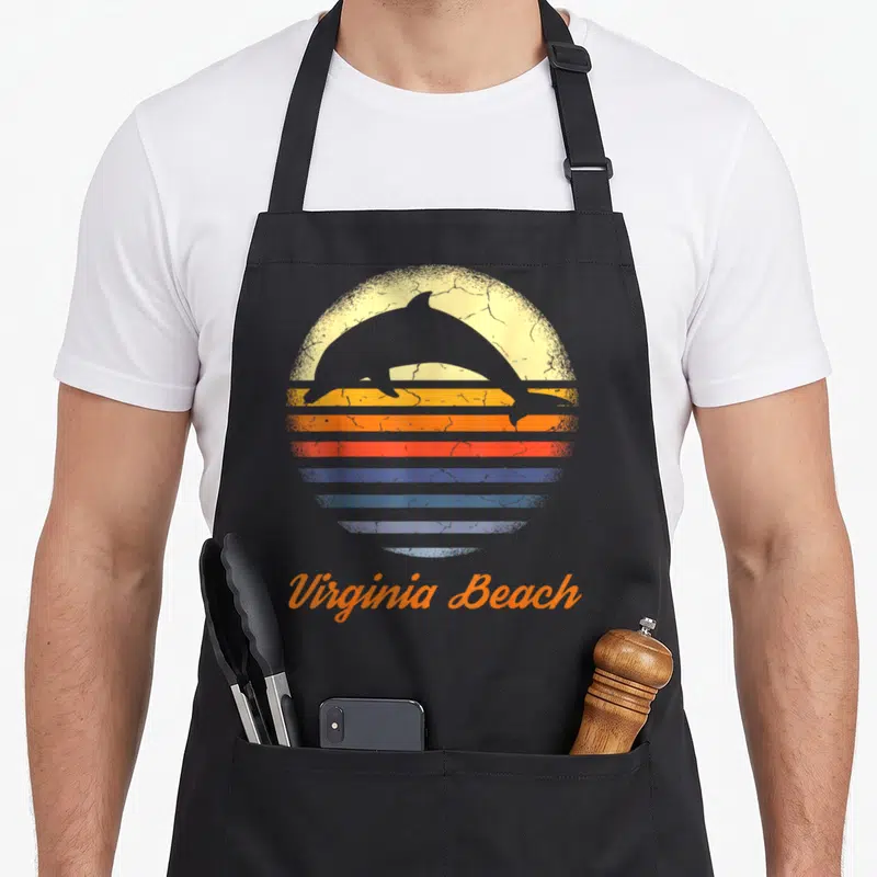 Virginia beach lover apron - Retro sunset with leaping dolphin silhouette and curved Virginia Beach text in orange — shown on a black cotton canvas apron against a white background