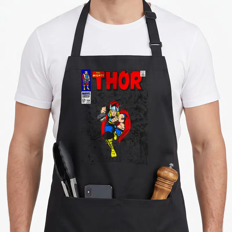 Mighty hobby apron - Retro Thor comic cover with red title text, leaping hero in blue and yellow, grey background crowd — shown on a black cotton canvas apron against a white background