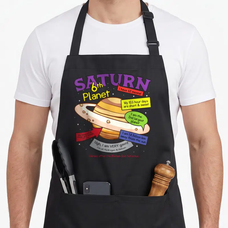 Planet teacher apron - Cartoon Saturn planet with purple title and colored fact bubbles listing moons and rings — shown on a black cotton canvas apron against a white background