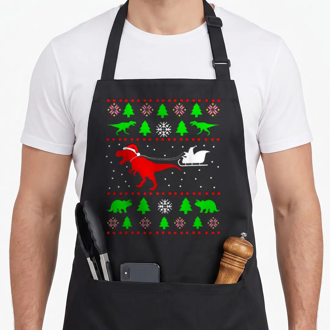 Dinosaur ugly christmas sweater apron - Red T-Rex pulling sleigh with pixelated green trees and white snowflakes — shown on a black cotton canvas apron against a white background