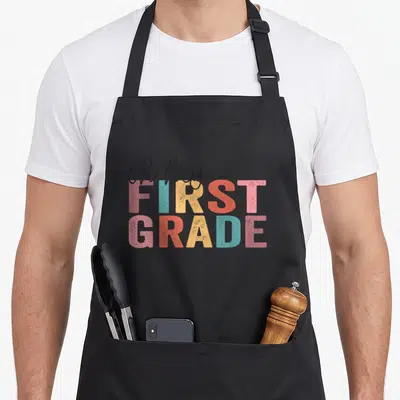 Hey grade teacher apron - Stacked oh hey FIRST GRADE text in pink blue yellow and red with distressed texture — shown on a black cotton canvas apron against a white background