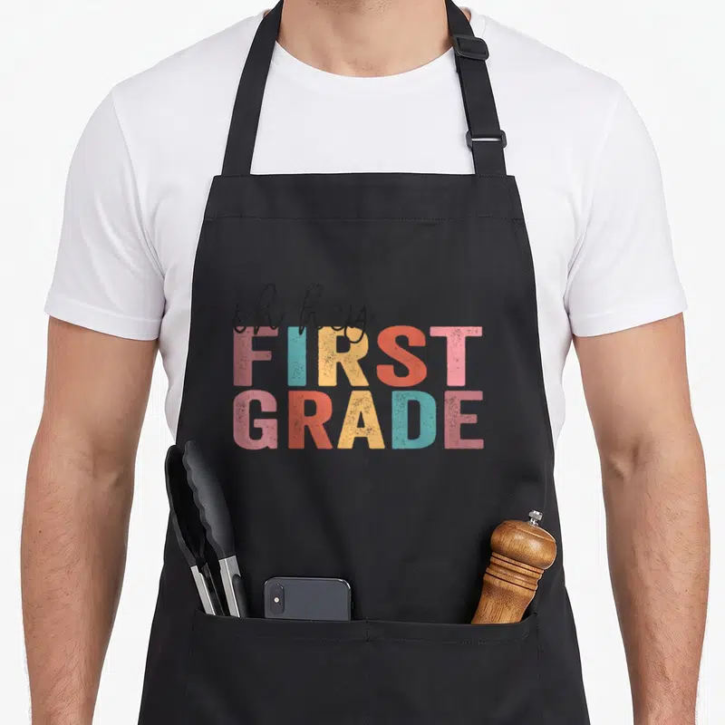 Hey grade teacher apron - Stacked oh hey FIRST GRADE text in pink blue yellow and red with distressed texture — shown on a black cotton canvas apron against a white background