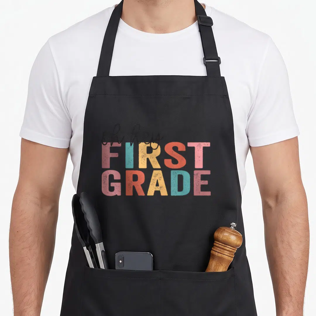 Hey grade teacher apron - Stacked oh hey FIRST GRADE text in pink blue yellow and red with distressed texture — shown on a black cotton canvas apron against a white background