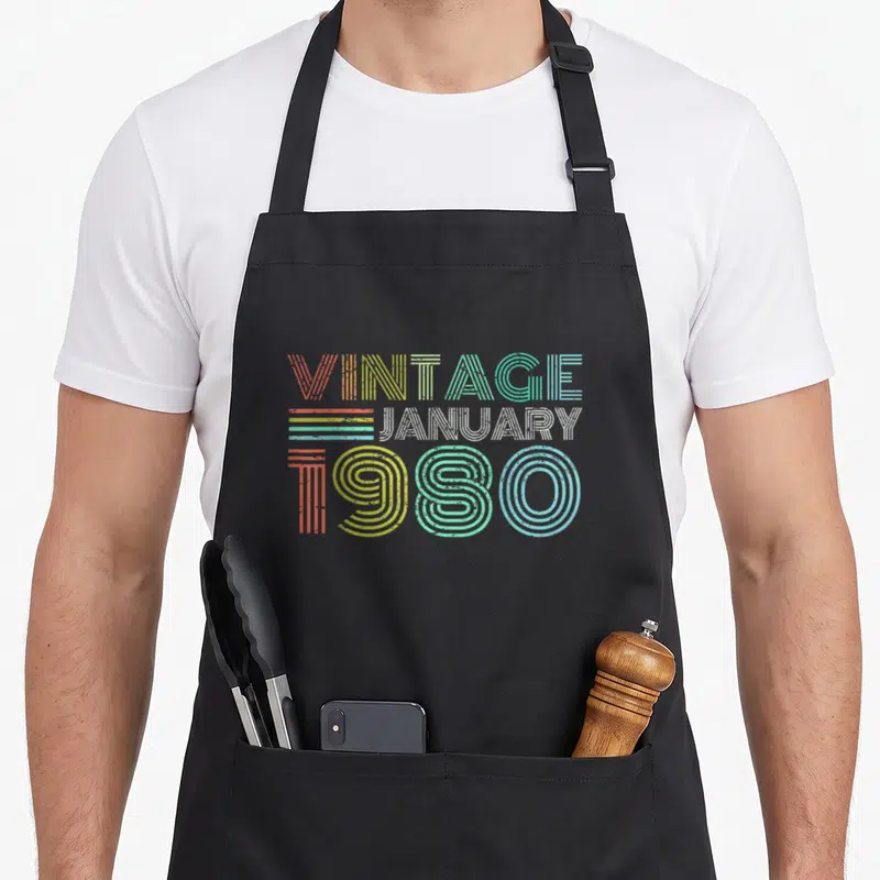 January forty 40th birthday apron - Stacked VINTAGE JANUARY 1980 text in retro orange, yellow, green, blue, and white — shown on a black cotton canvas apron against a white background