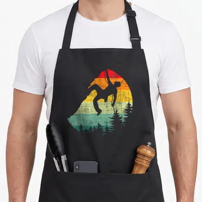 Mountain climber bouldering apron - Grey climber silhouette over red orange yellow teal striped sun with pine trees — shown on a black cotton canvas apron against a white background