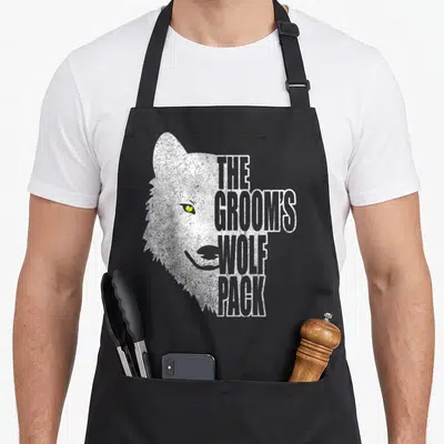 Groom police squad apron - Distressed wolf head with yellow eye and stacked text reading The Groom's Wolf Pack — shown on a black cotton canvas apron against a white background