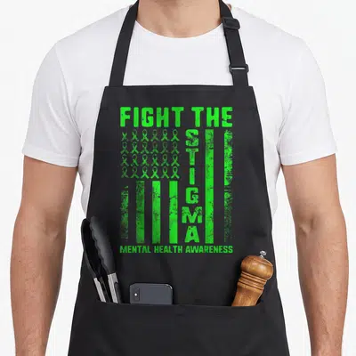 Stigma anxiety apron - Green awareness ribbons arranged in a grid with vertical STIGMA text and distressed typography — shown on a black cotton canvas apron against a white background