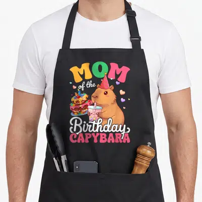 Birthday cartoon apron - MOM of the Birthday CAPYBARA text with capybara wearing party hat holding drink next to cake — shown on a black cotton canvas apron against a white background