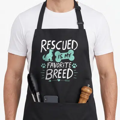 Saved dog breed aprons - Rescued Is My Favorite Breed text with mint dog, bone, paw prints and heart arrow — shown on a black cotton canvas apron against a white background