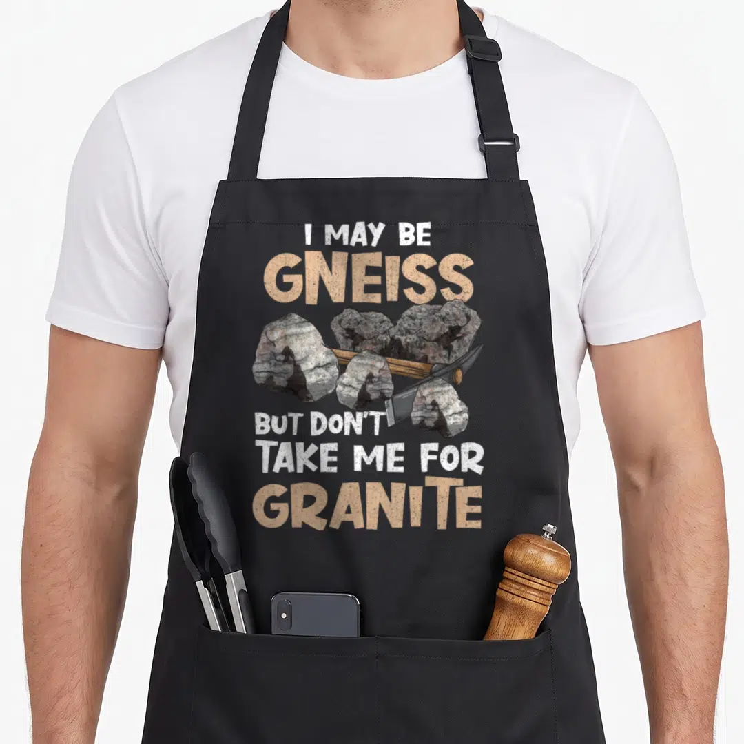 Geologist science apron - I may be gneiss but don't take me for granite text over grey rocks and hammer — shown on a black cotton canvas apron against a white background