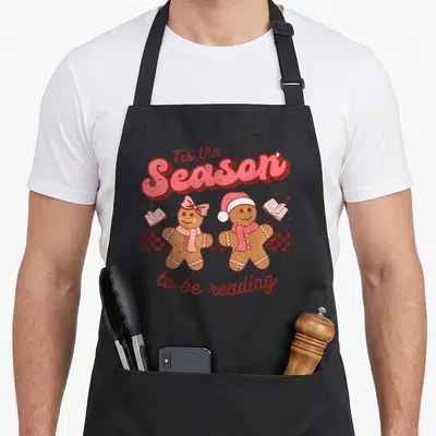 School christmas apron - Two gingerbread people with open books and Tis the Season to be reading text in pink and red — shown on a black cotton canvas apron against a white background