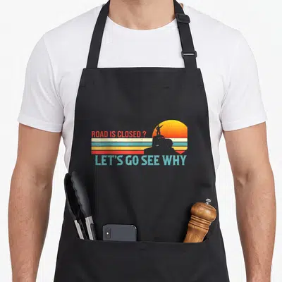 Closed road apron - Retro sun graphic with vehicle silhouette and text ROAD IS CLOSED LET'S GO SEE WHY — shown on a black cotton canvas apron against a white background