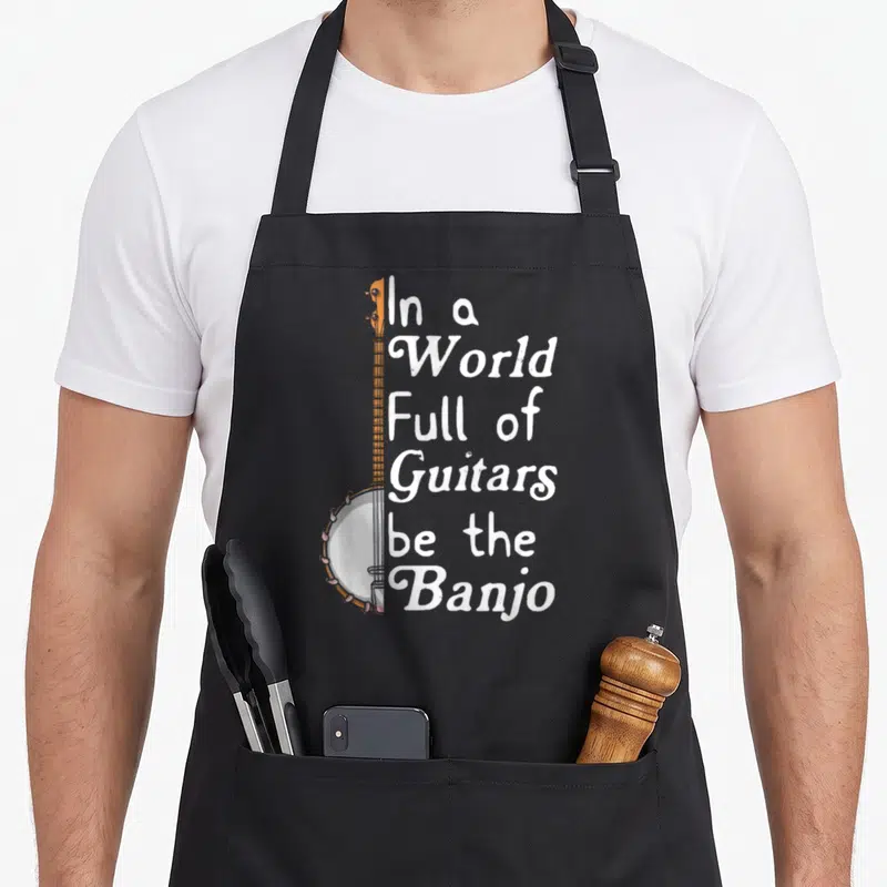 Banjo bluebird aprons - Vertical banjo overlapping stacked white text reading In a World Full of Guitars be the Banjo — shown on a black cotton canvas apron against a white background
