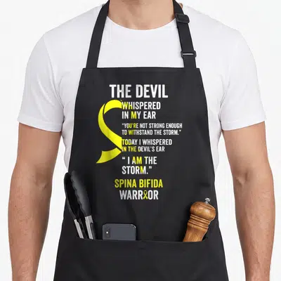 Spina devil's apron - Yellow awareness ribbon and white text reading Spina Bifida Warrior with storm quote — shown on a black cotton canvas apron against a white background