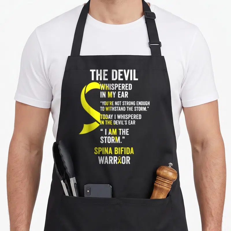 Spina devil's apron - Yellow awareness ribbon and white text reading Spina Bifida Warrior with storm quote — shown on a black cotton canvas apron against a white background