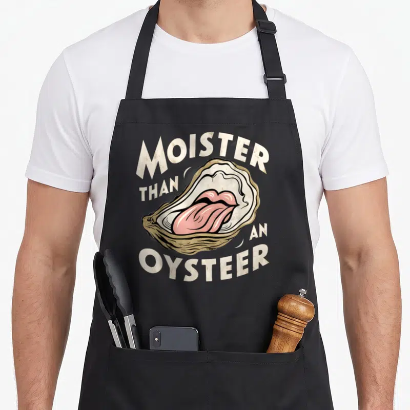 Moisture oyster apron - Cartoon open oyster with pink tongue and arched text reading MOISTER THAN AN OYSTEER — shown on a black cotton canvas apron against a white background