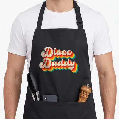 Disco daddy apron - Retro stacked typography reading Disco Daddy with orange yellow and blue shadows — shown on a black cotton canvas apron against a white background