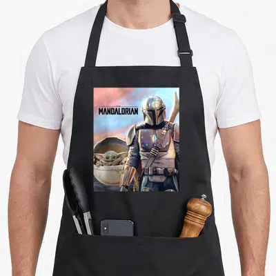 Adventure ivorian apron - The Mandalorian text with armored figure and Grogu in pod using silver brown blue pink — shown on a black cotton canvas apron against a white background