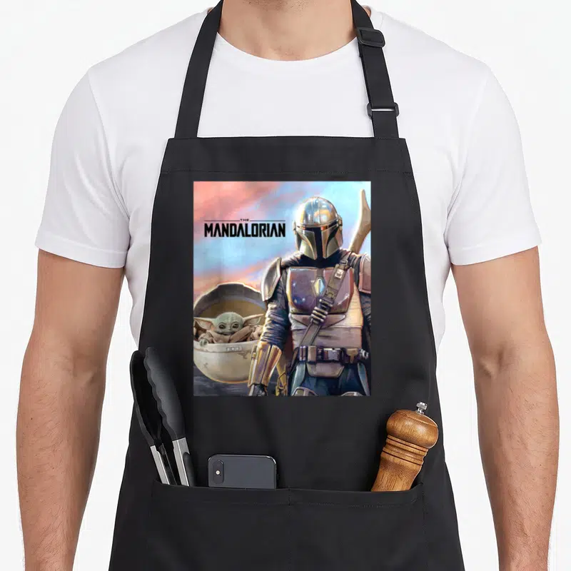 Adventure ivorian apron - The Mandalorian text with armored figure and Grogu in pod using silver brown blue pink — shown on a black cotton canvas apron against a white background