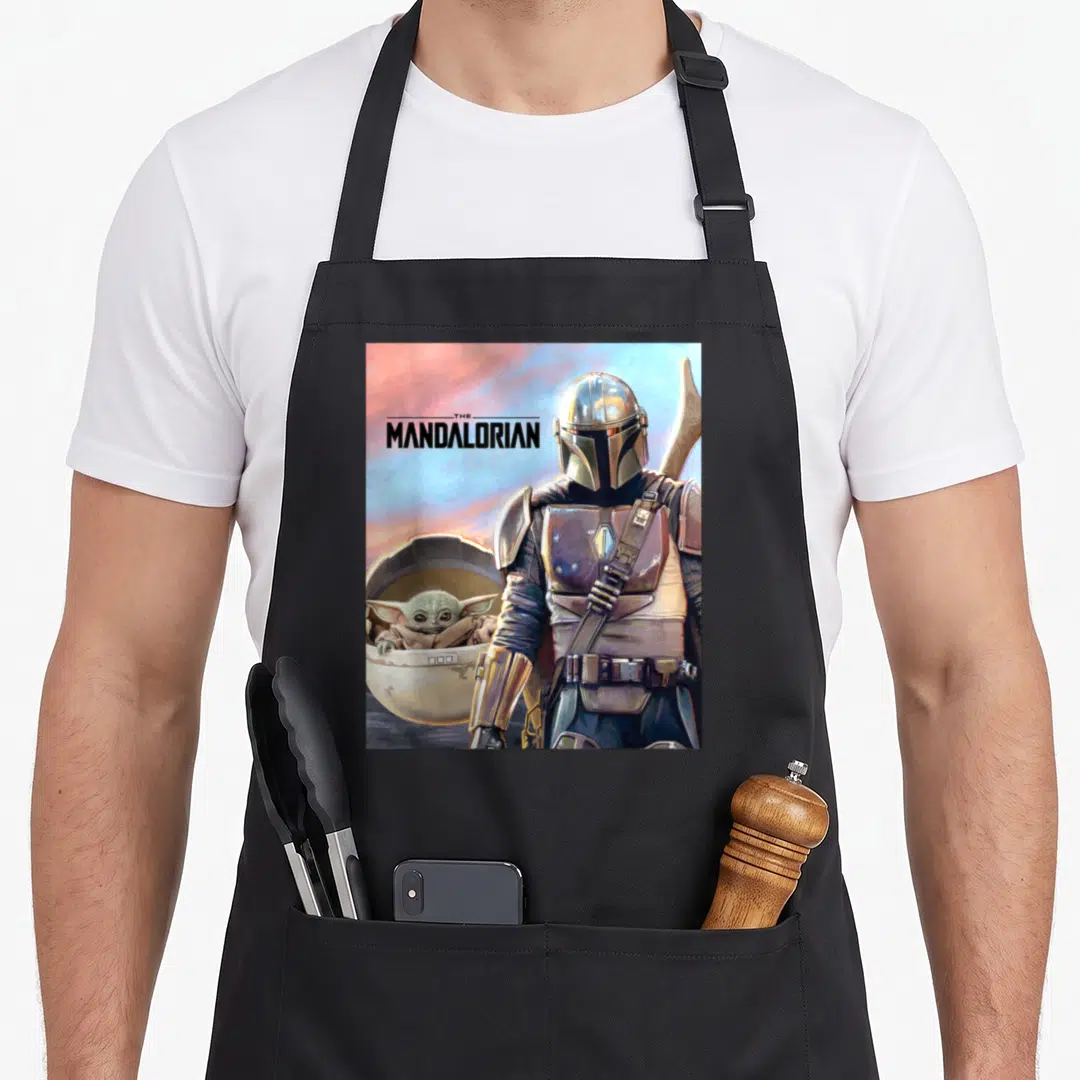 Adventure ivorian apron - The Mandalorian text with armored figure and Grogu in pod using silver brown blue pink — shown on a black cotton canvas apron against a white background