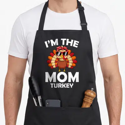 Family thanksgiving funny turkey apron - Cartoon turkey with red sunglasses and polka dot headband between I'M THE MOM — shown on a black cotton canvas apron against a white background