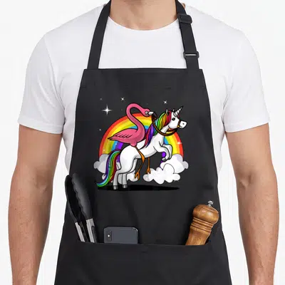 Unicorn flamingo apron - Pink flamingo riding white unicorn with rainbow mane over rainbow arch and stars — shown on a black cotton canvas apron against a white background