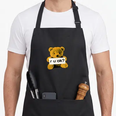 Yungblud r aprons - Orange cartoon teddy bear holding white sign with r u ok? text in black — shown on a black cotton canvas apron against a white background